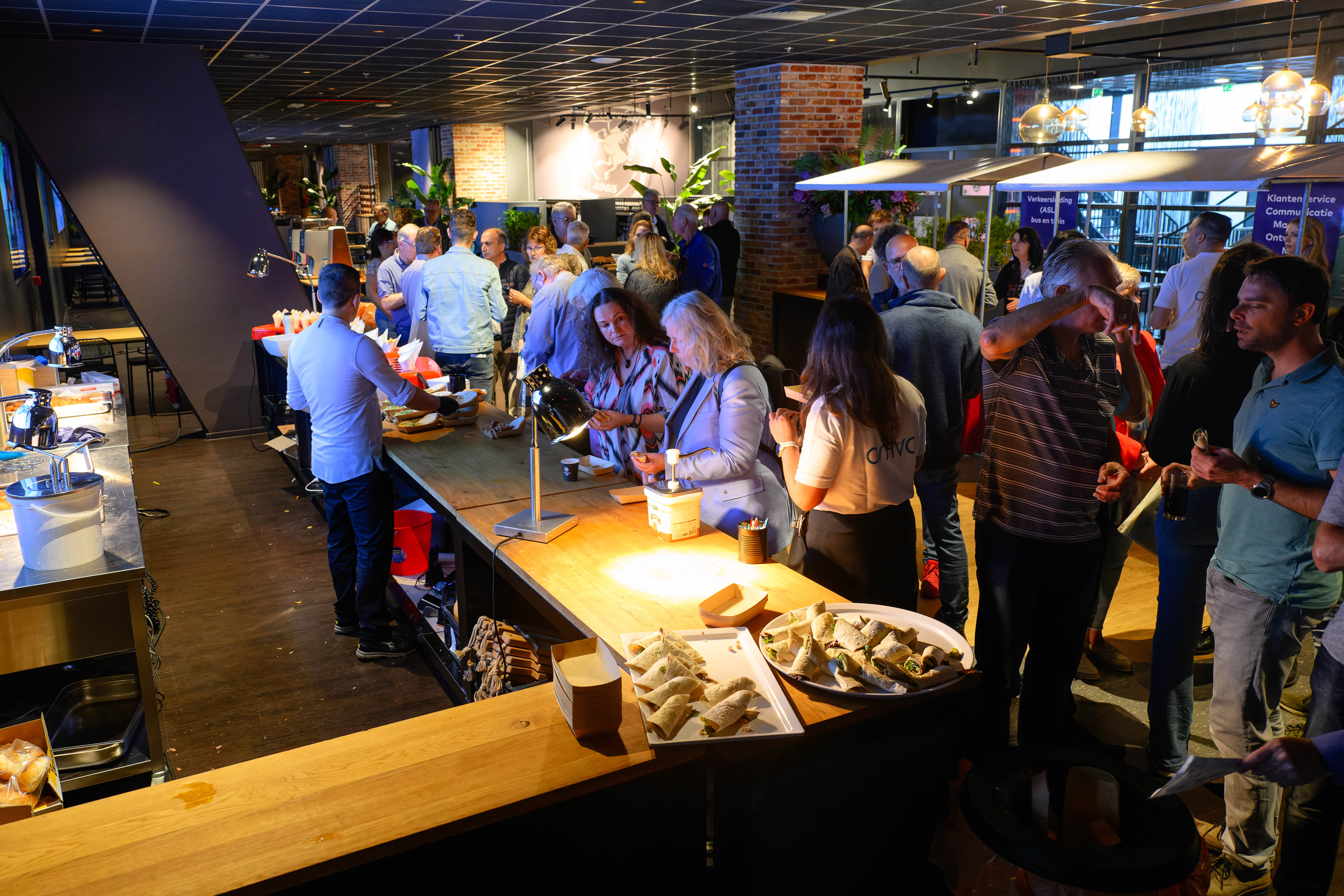 Groep mensen staat in een ruimte met stands en een bar.