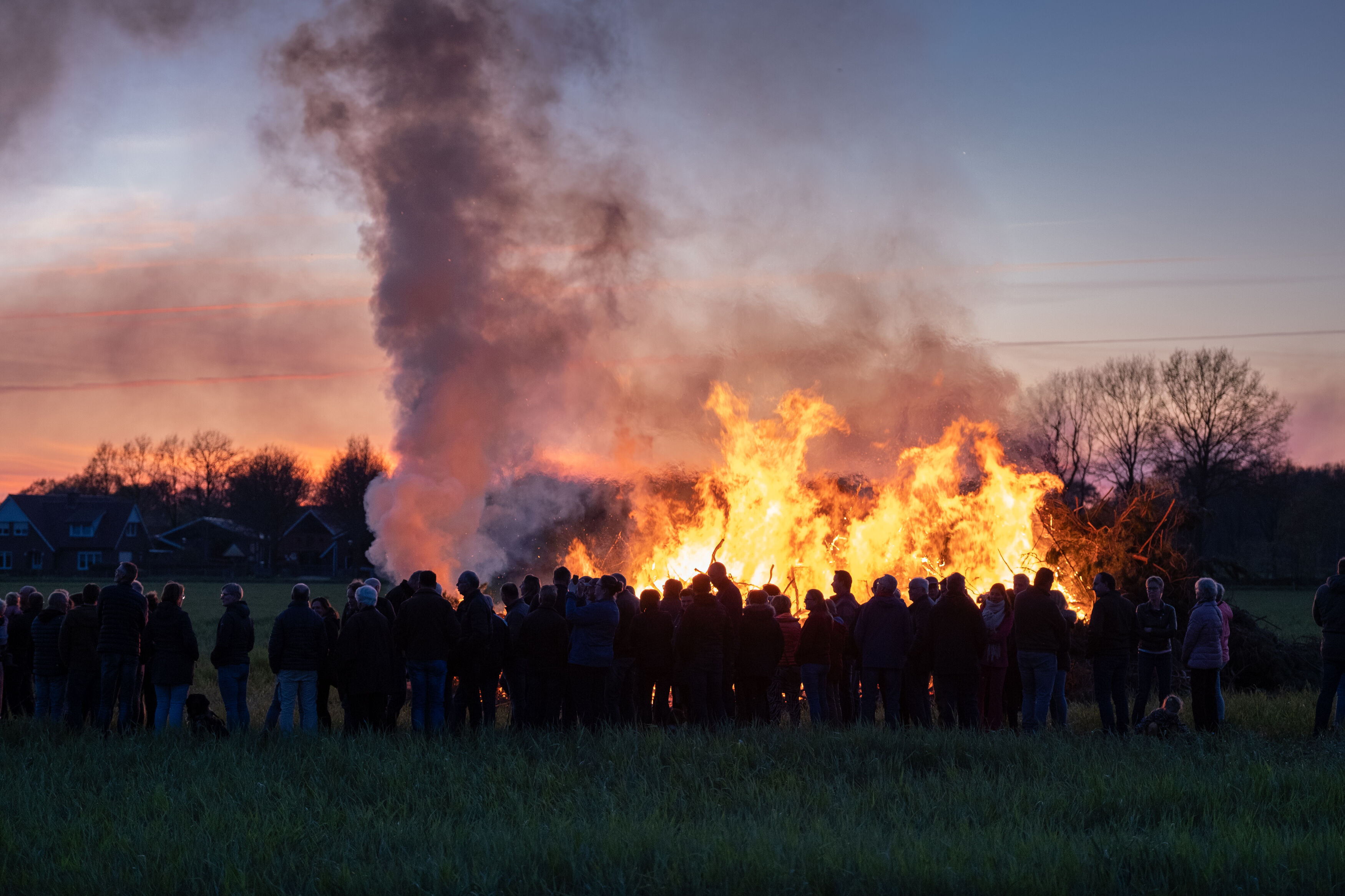 een schemeravond waar je een aangestoken paasvuur ziet met rook en een groep mensen om het vuur heen