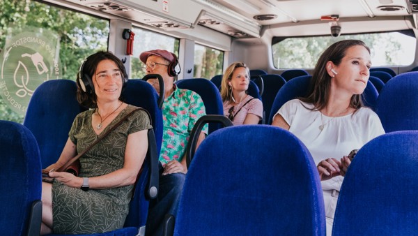 Vier mensen zitten in een bus van Arriva op blauwe stoelen. Ze hebben koptelefoons op of in en kijken geïnteresseerd naar buiten.