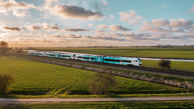 Een blauw-witte Arriva-trein rijdt over het spoor tussen weilanden met een ondergaande zon en een prachtige wolkenlucht.