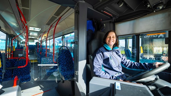 Een vrouwelijke buschauffeur met bruin haar zit achter het stuur van een Arriva-bus en glimlacht.