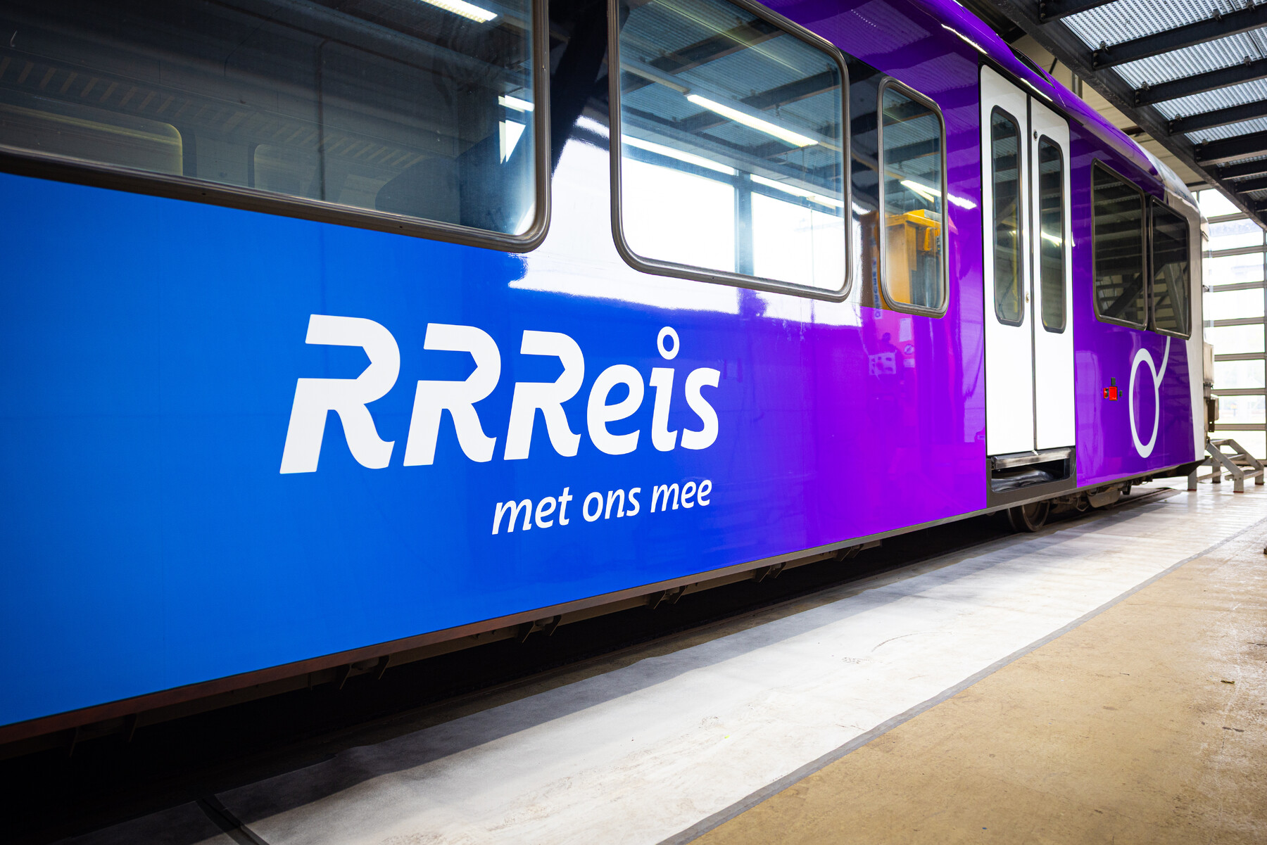 Bestickering van een RRReis trein in Achterhoek en Rivierenland, logo van RRReis met tekst: RRReis met ons mee. Trein in een werkplaats met blauwe en paarse huisstijlkleuren.