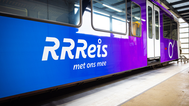 RRReis trein in Achterhoek en Rivierenland Bestickering van een RRReis trein in Achterhoek en Rivierenland, logo van RRReis met tekst: RRReis met ons mee. Trein in een werkplaats met blauwe en paarse huisstijlkleuren.