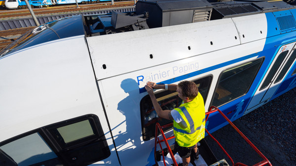 Een man in een geel hesje plakt de letters 'Reinier Paping' op een blauw-witte Arriva-trein.