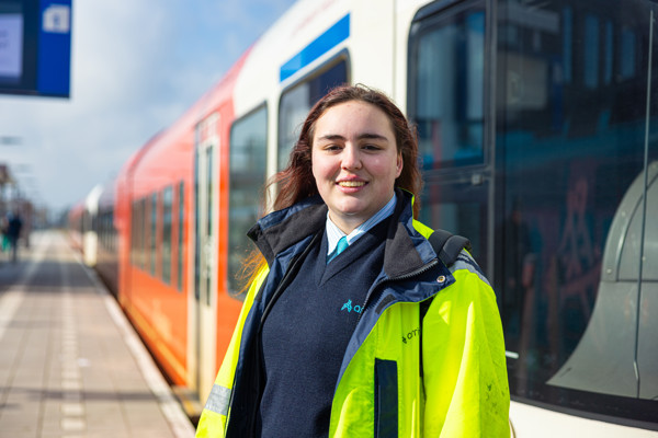Een vrouwelijke machinist in uniform staat voor een rood-witte Arriva-trein en lacht. De zon schijnt.