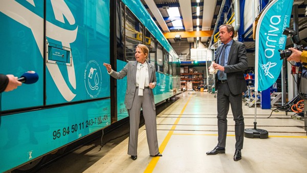Een vrouw en een man in nette kleding worden geïnterviewd terwijl ze naar een blauwe Arriva-trein in een werkplaats kijken.