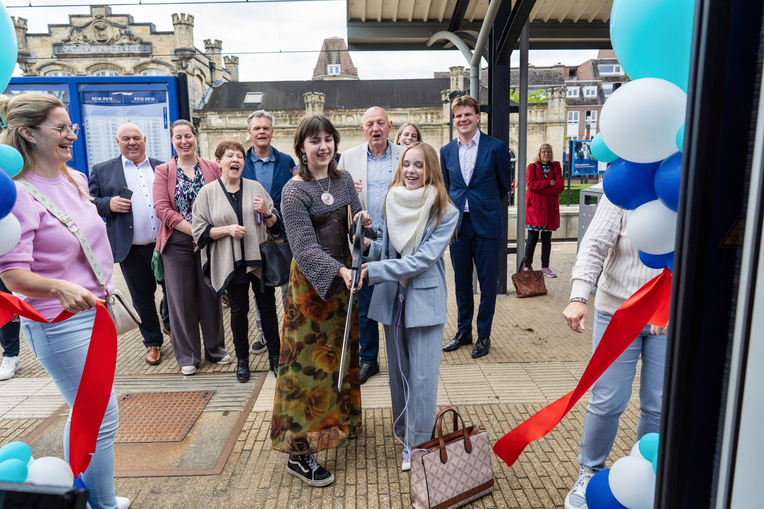 op het perron in Valkenburg staat een vrolijk lachende groep mensen. In het midden staan zangeres Emma Kok en woordkunstenaar Eline Willaert en knippen officieel het lint  door