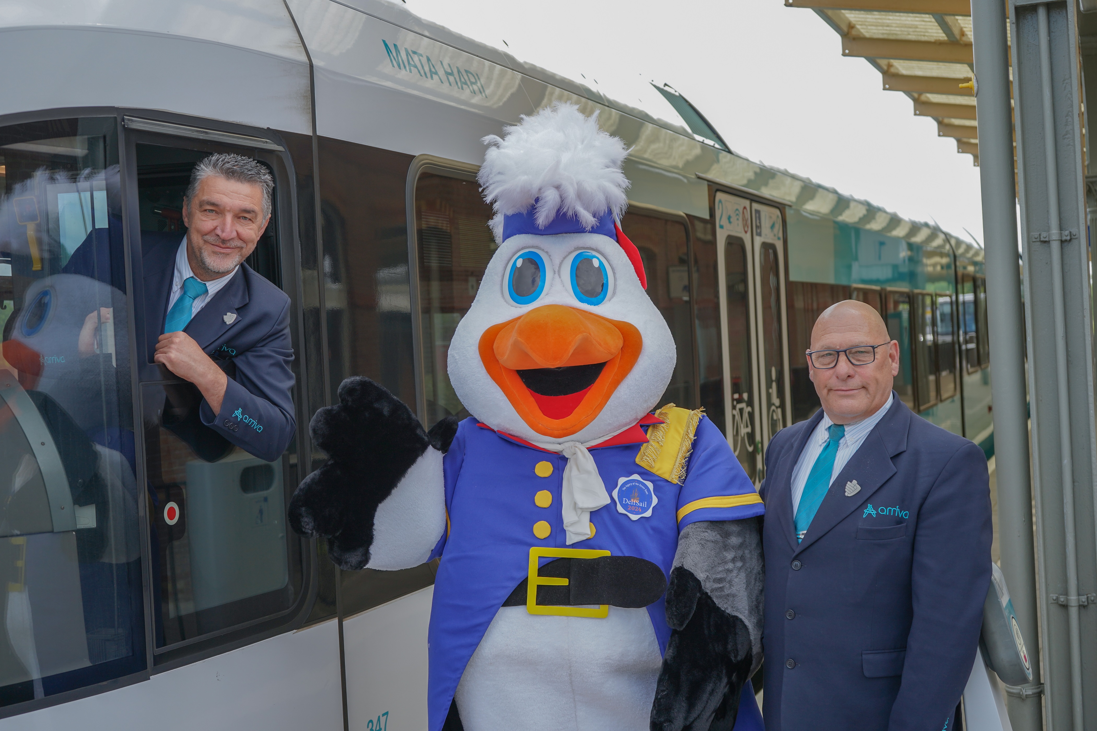 machinist hangt met zijn hoofd en een arm uit het raam van een stilstaande trein. Op het perron naast de machinist staat een steward samen met de mascotte van Delfsail.