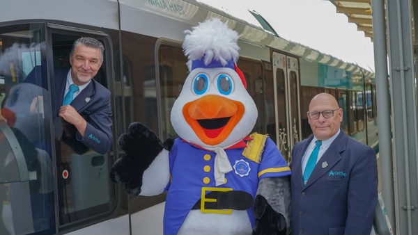 machinist hangt met zijn hoofd en een arm uit het raam van een stilstaande trein. Op het perron naast de machinist staat een steward samen met de mascotte van Delfsail. machinist hangt met zijn hoofd en een arm uit het raam van een stilstaande trein. Op het perron naast de machinist staat een steward samen met de mascotte van Delfsail.