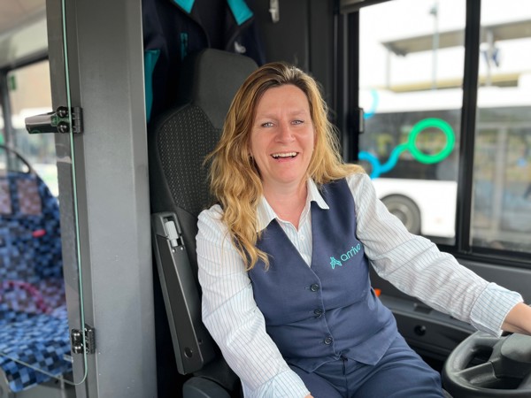 Een vrouwelijke buschauffeur met lang blond haar zit achter het stuur van een Arriva-bus en lacht.