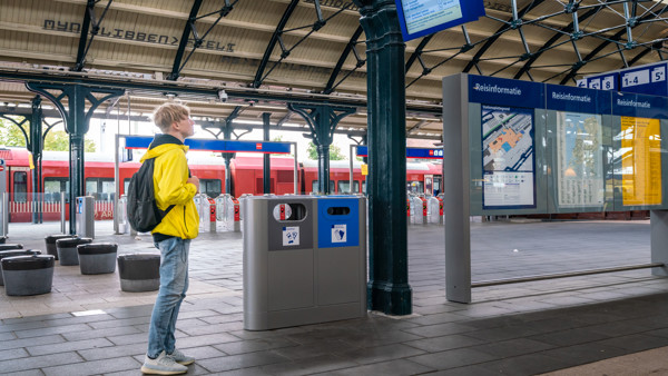 Tiener in felgele jas staat naar de vertrektijden te kijken op een treinperron. Op de achtergrond staat een rode Arriva-trein. Tiener in felgele jas staat naar de vertrektijden te kijken op een treinperron. Op de achtergrond staat een rode Arriva-trein.