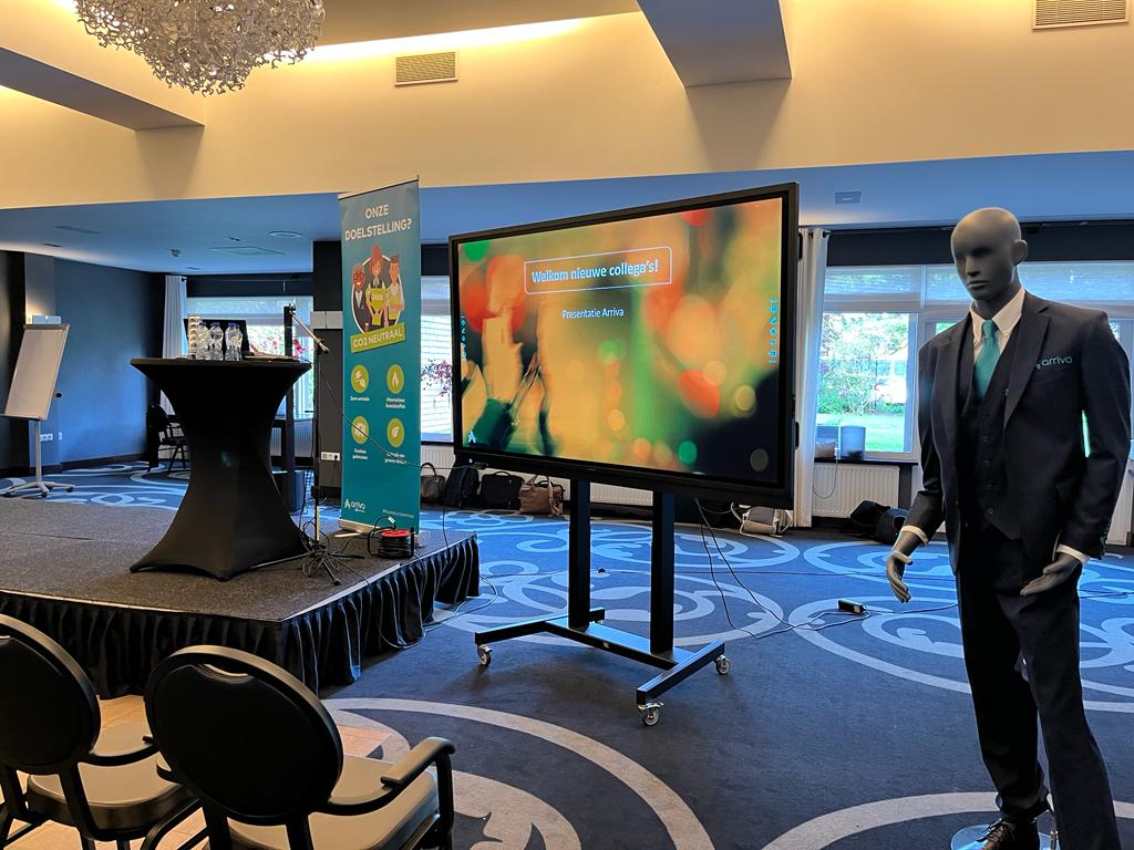 Een nog lege zaal met podium, presentatiescherm, stoelen en paspop in Arriva-uniform.