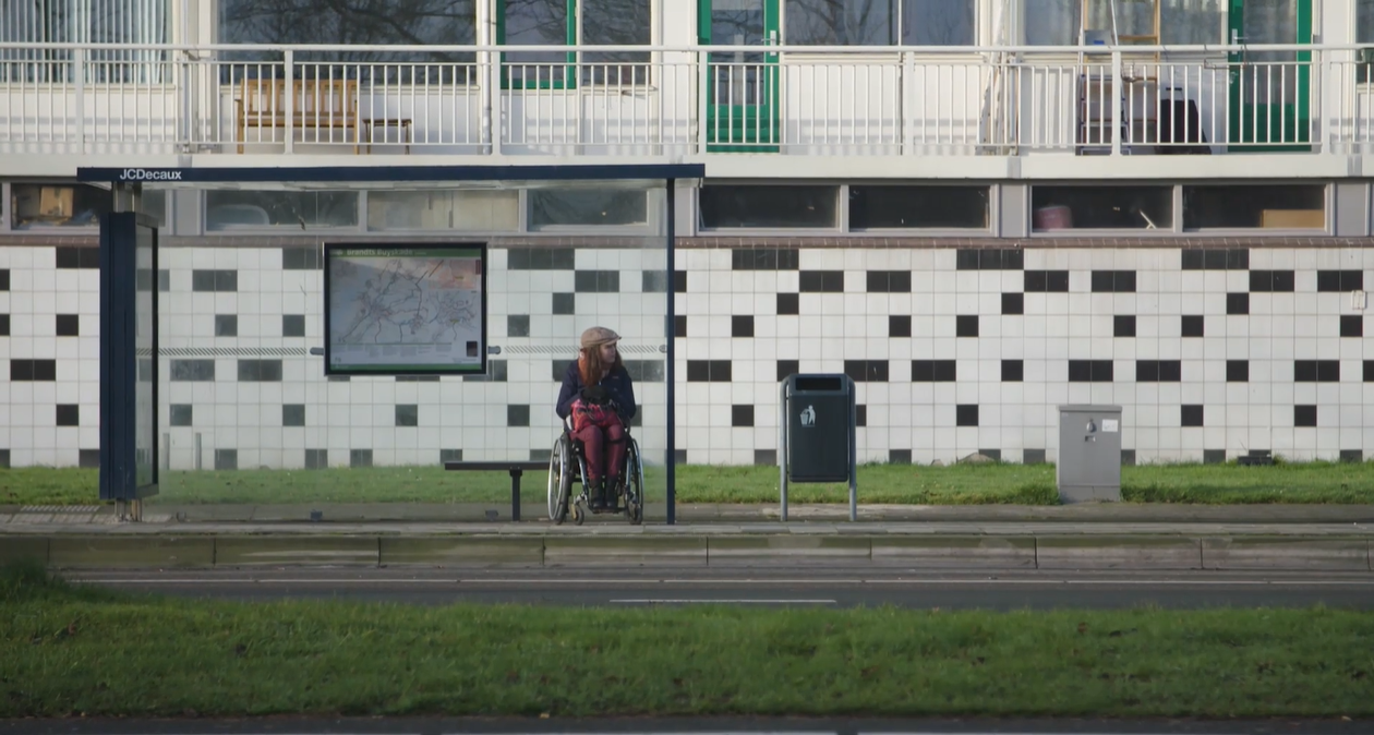 Een vrouw in een rolstoel zit bij een bushokje voor een gebouw.