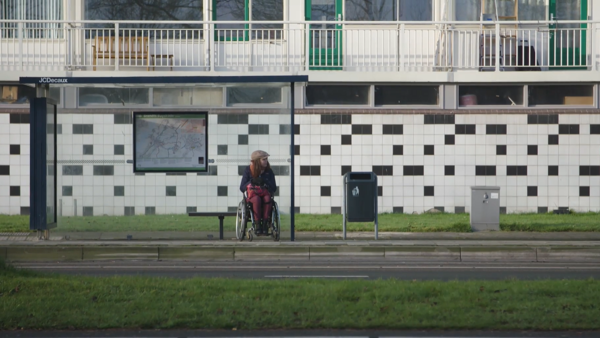 Een vrouw in een rolstoel zit bij een bushokje voor een gebouw.