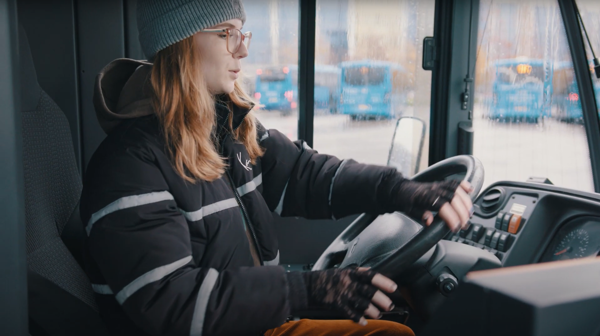  Een vrouwelijke buschauffeur met rood haar en een muts zit achter het stuur en bestuurt de bus.