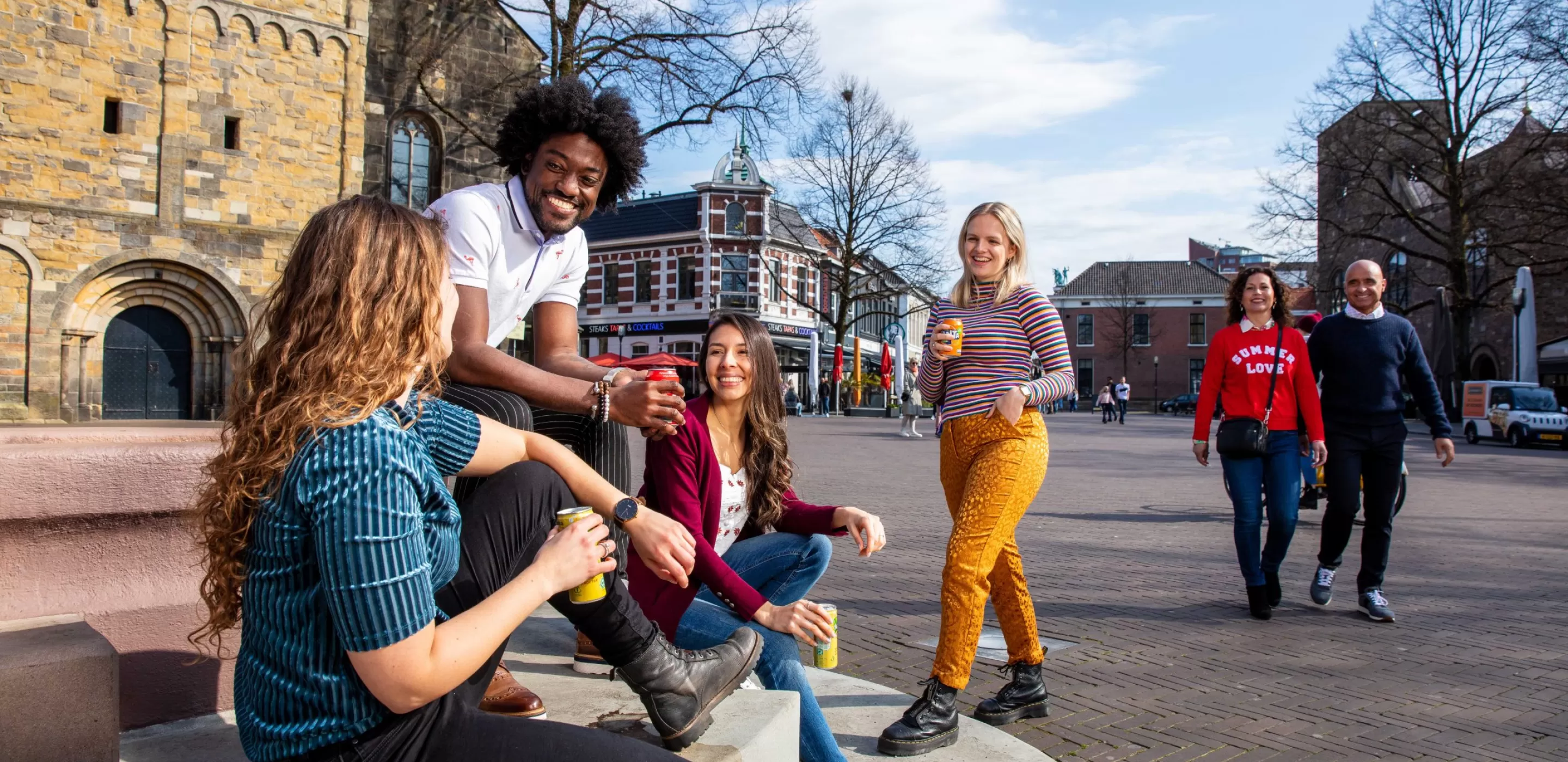 Op een oud stadsplein zitten 3 mensen op een stoepje en lachen met elkaar. Er komt een meisje lachend naar ze toe gelopen. Op de achtergrond zie je een echtpaar samen voorbij komen.