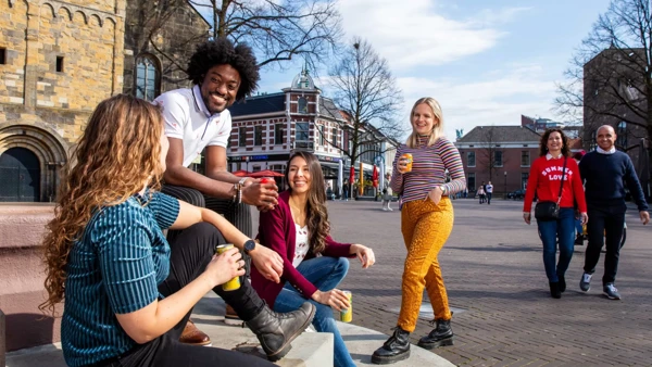 Op een oud stadsplein zitten 3 mensen op een stoepje en lachen met elkaar. Er komt een meisje lachend naar ze toe gelopen. Op de achtergrond zie je een echtpaar samen voorbij komen. Op een oud stadsplein zitten 3 mensen op een stoepje en lachen met elkaar. Er komt een meisje lachend naar ze toe gelopen. Op de achtergrond zie je een echtpaar samen voorbij komen.