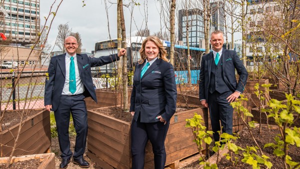 Drie mensen in Arriva-uniform staan tussen plantenbakken met bomen en lachen naar de camera.