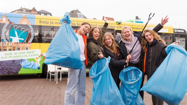 Een groep jonge meiden poseert voor de camera met afval in hun handen, met een Arriva-bus op de achtergrond.