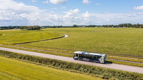 Een blauw-witte Arriva-bus rijdt tussen de weilanden, gezien vanuit een vogelperspectief.