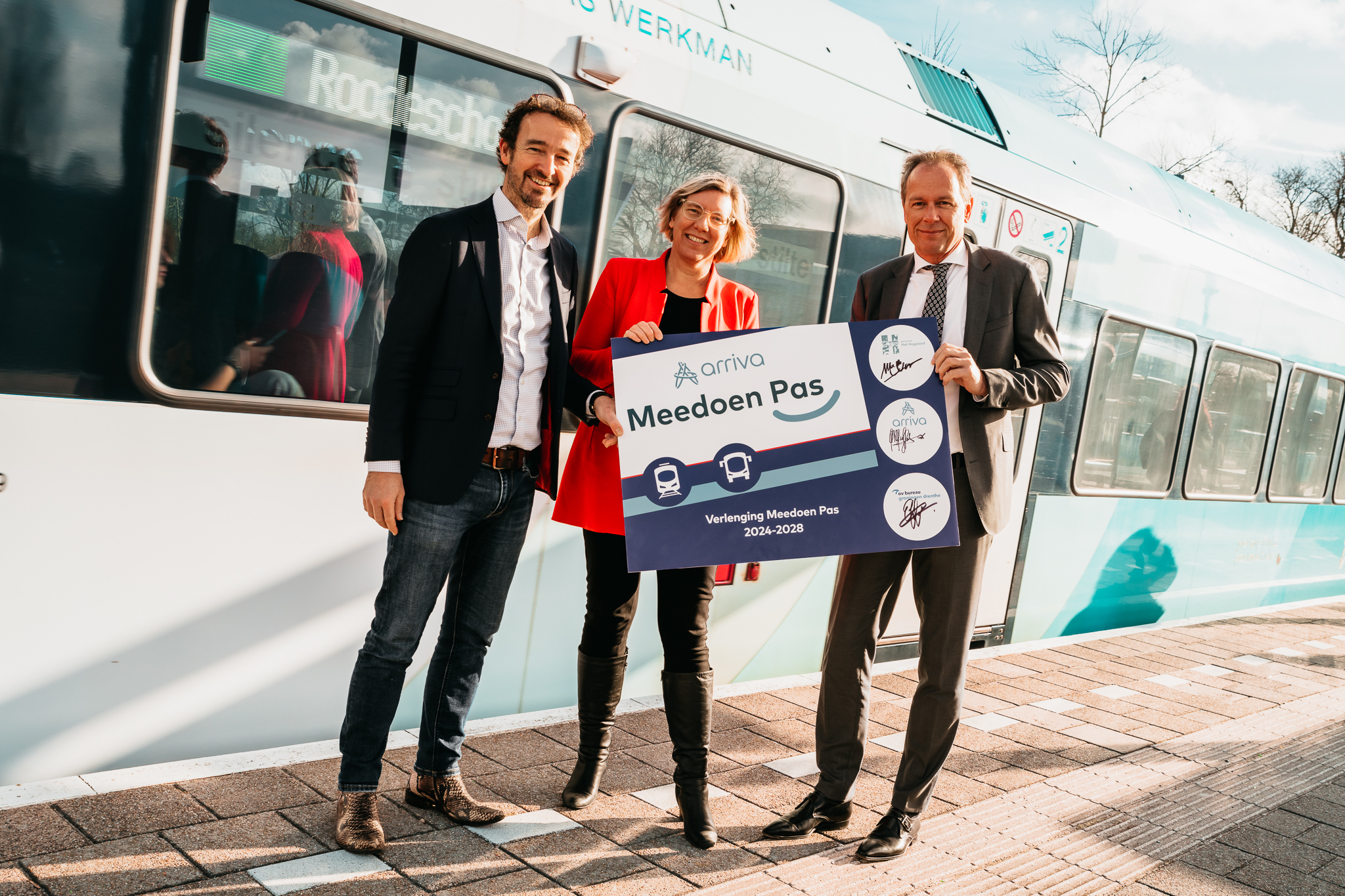 drie mensen op het station met een vergrootte meedoen pas in de hand met op de achtergrond een arriva trein