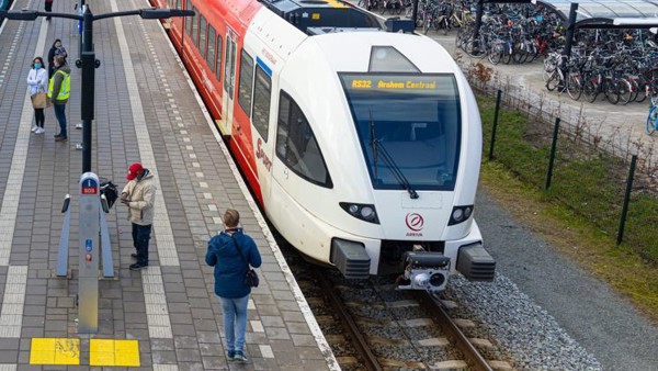 Een bovenaanzicht van een rood-witte Arriva-trein langs het perron, met in de verte een fietsenstalling en gebouwen.