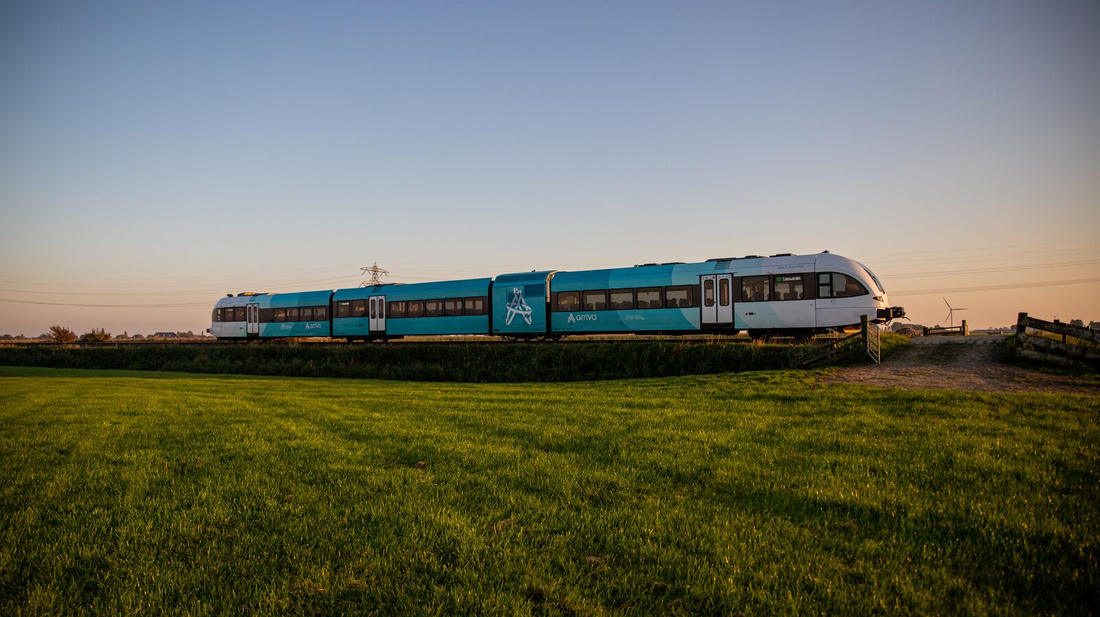 Een blauwe Arriva-trein rijdt door een groen landschap onder een strakblauwe lucht vlak voor zonsopkomst.