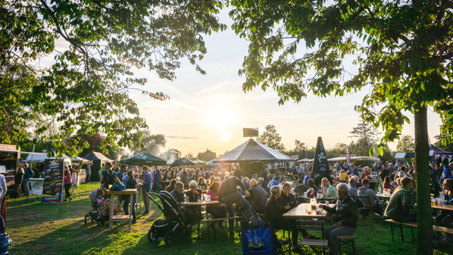 Drukbezocht buitenfestival bij zonsondergang, met mensen aan picknicktafels op een grasveld omringd door bomen. Drukbezocht buitenfestival bij zonsondergang, met mensen aan picknicktafels op een grasveld omringd door bomen.