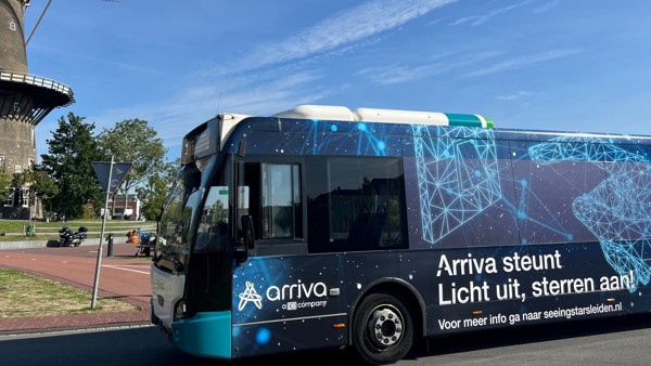 Een Arriva-bus met een afbeelding van het heelal en de tekst "Arriva steunt. Licht uit, sterren aan." rijdt langs een molen.