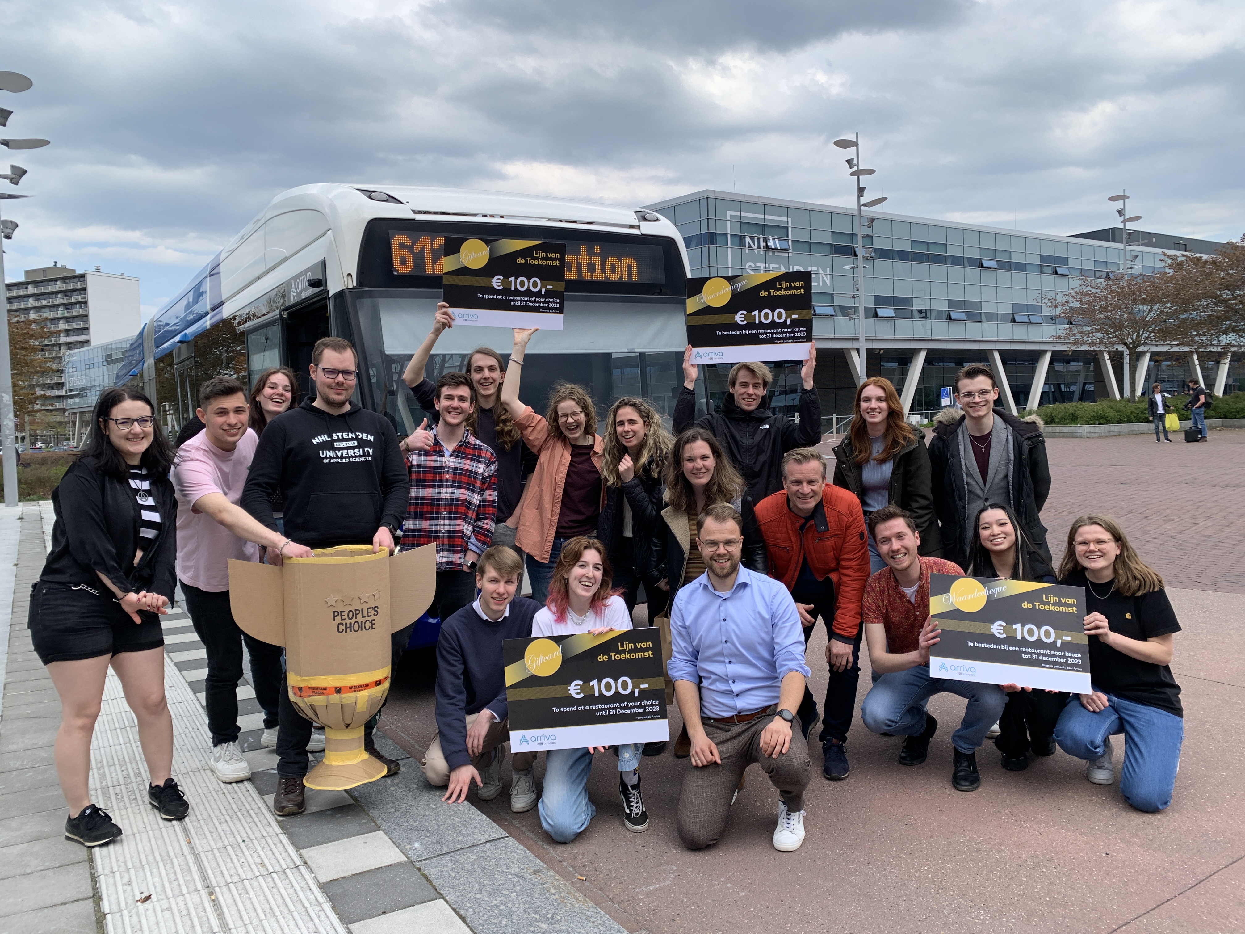 Groep mensen poseert lachend voor een Arriva-bus terwijl ze giftcards van 'Lijn van de toekomst' vasthouden.