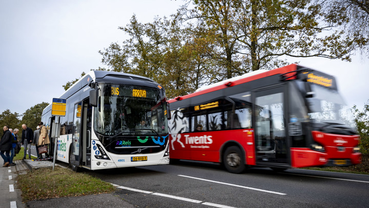 Een witte Arriva-bus met Rrreis erop staat bij de halte en wordt ingehaald door een rode Arriva-bus.