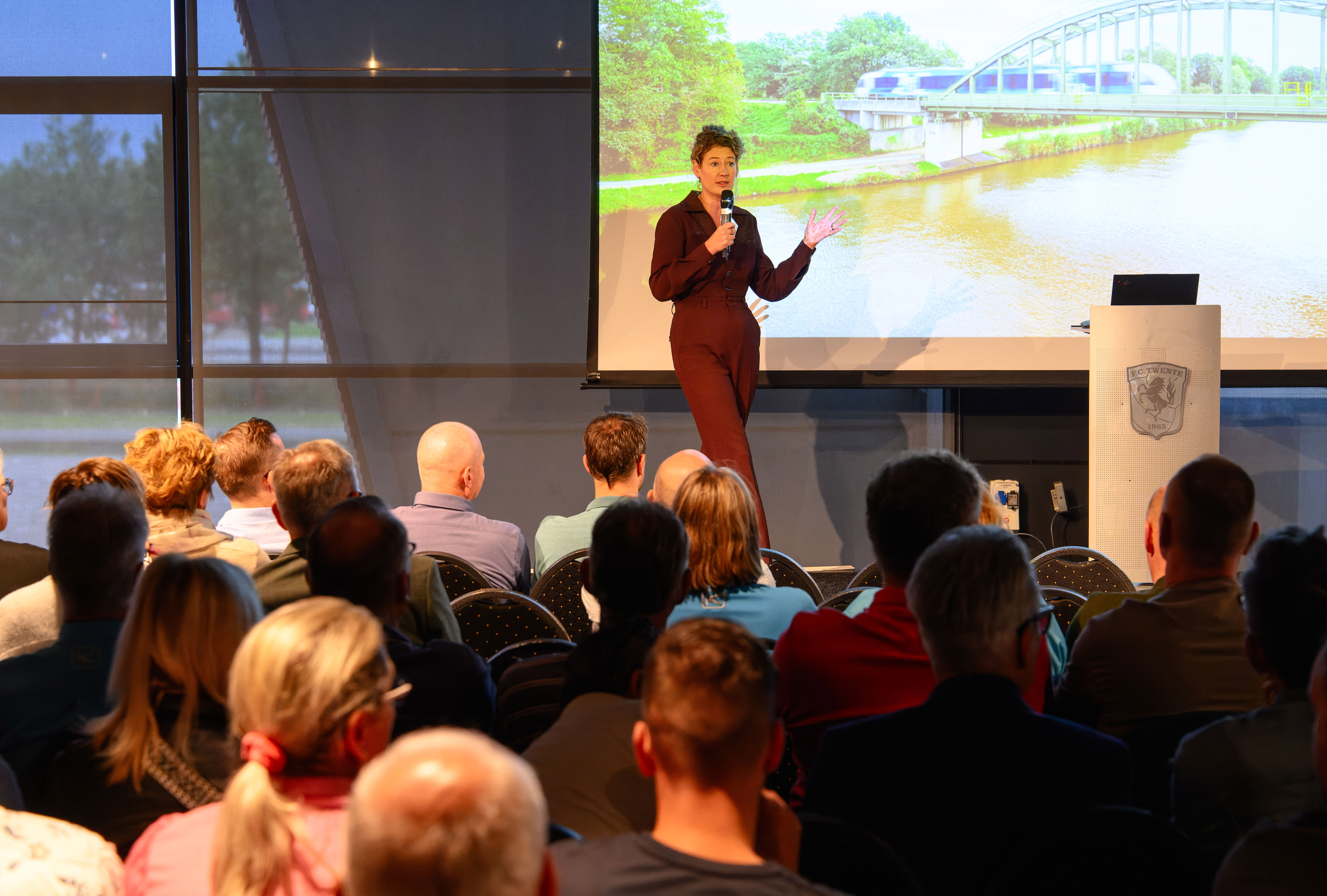 Vrouw in rood broekpak speecht voor groot gezelschap, achter haar een projectiescherm met daarop een trein van Arriva die over een spoorbrug rijdt.
