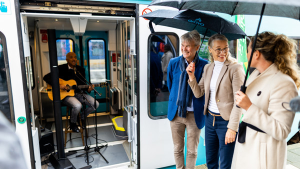 Een man speelt gitaar in een Arriva-trein terwijl de deur openstaat. Buiten de trein staan een paar mensen onder paraplu's.