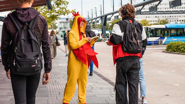 Iemand verkleed als een kip deelt flyers uit bij een busstation.