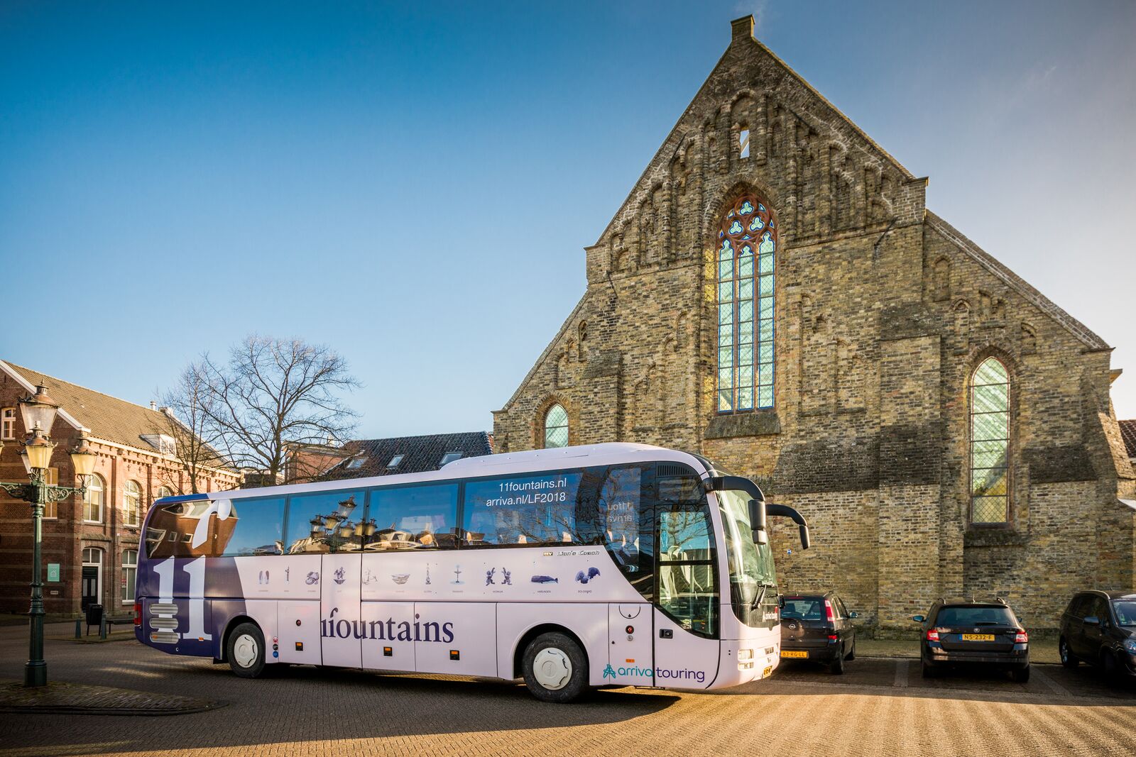 Een Arriva Touring-bus met reclame voor 11Fountains in Friesland staat voor een kerk in het zonlicht.