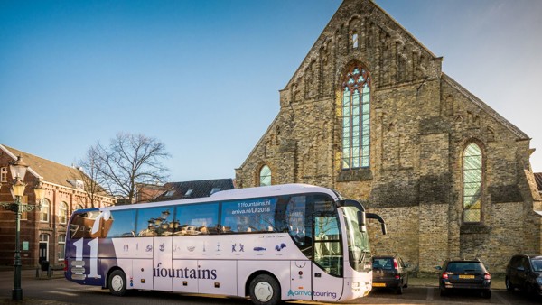 Een Arriva Touring-bus met reclame voor 11Fountains in Friesland staat voor een kerk in het zonlicht.
